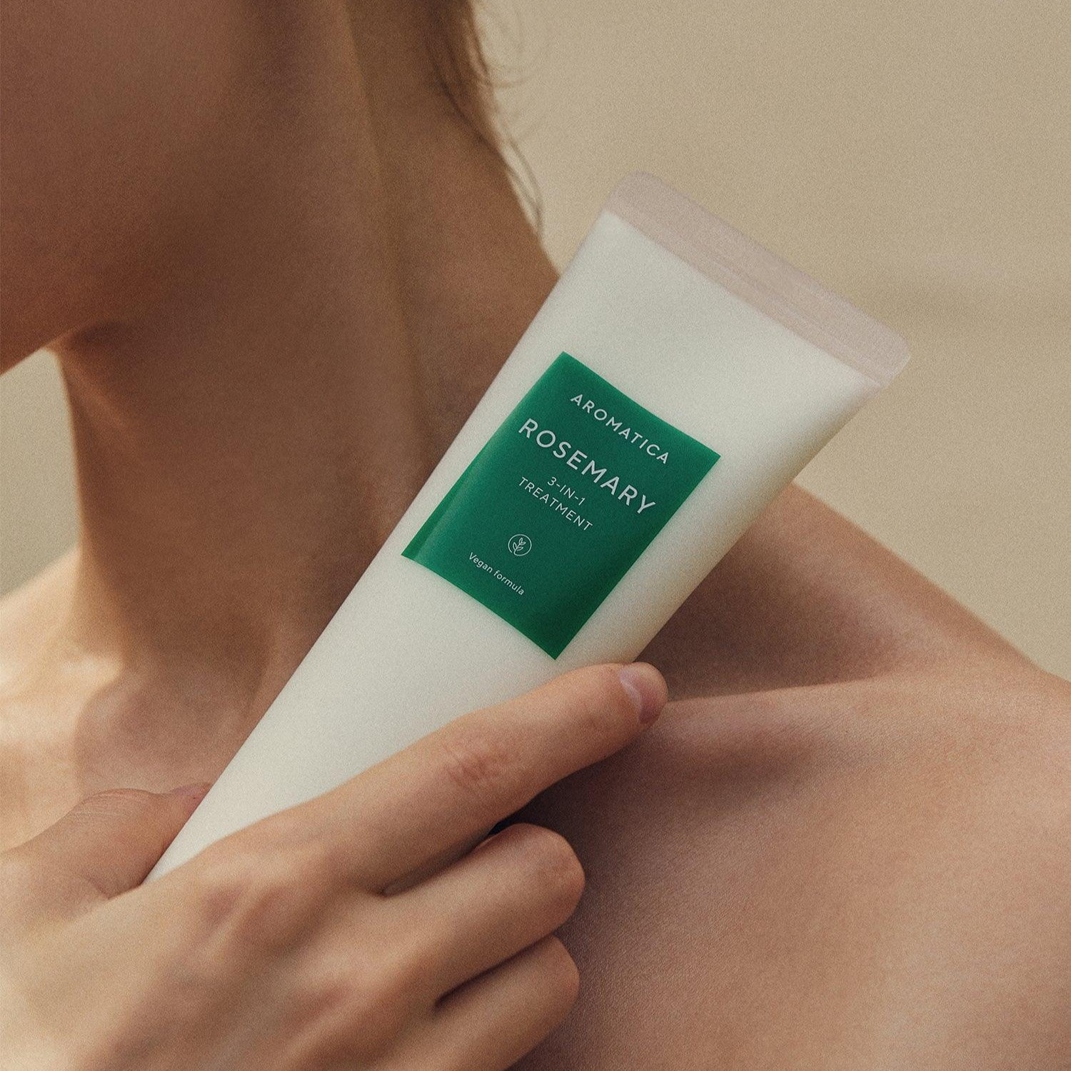 Hand holding a tube of Aromatica Rosemary hair treatment against a neutral background