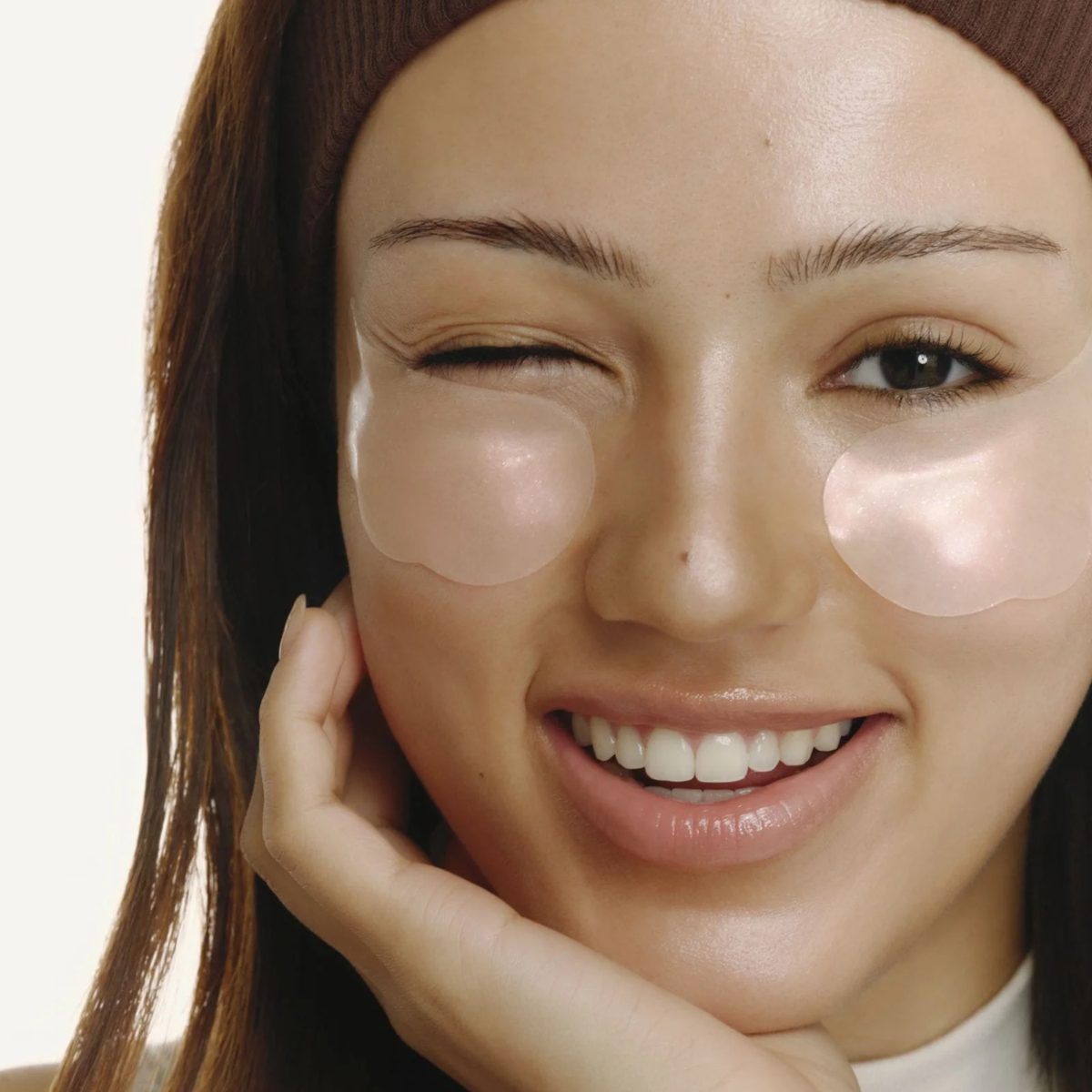 A close-up of a woman with clear under-eye patch gel applied to both eyes, wearing a brown beanie and smiling with one eye winking. The background is a clean, solid off-white.