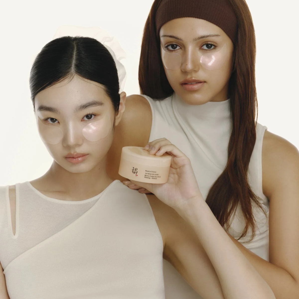 Two models wearing beige skincare sets hold a beige cosmetic jar against a clean off-white background.