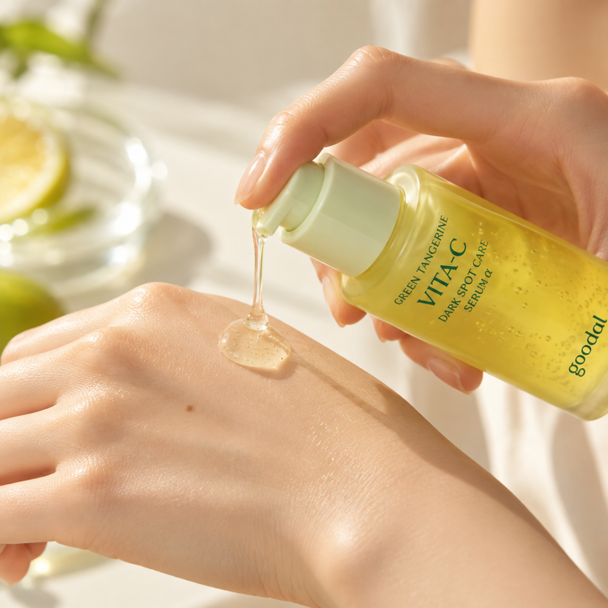 A hand pours a clear gel-like serum from a yellow bottle labeled VITA-C Green Peony Dual Spot Care onto the back of another hand, with a softly blurred background showing a glass and lime slices.