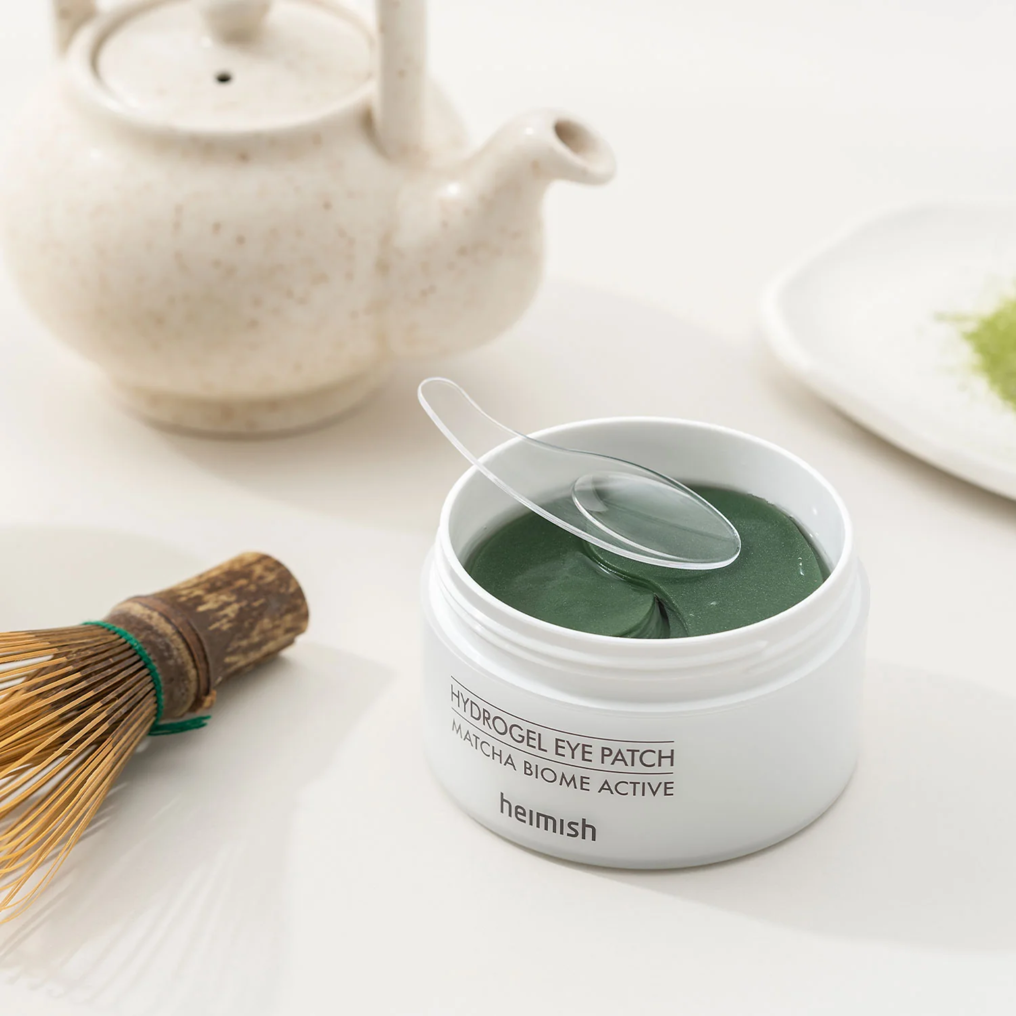 A white jar of Heimish Hydrogel Eye Patch with a clear plastic spoon resting inside, set on a white surface with a ceramic teapot and matcha whisk in the background.