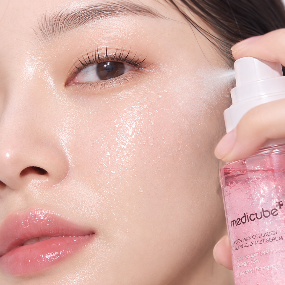 Close-up of a person's face holding and spraying a pink Medicebe glow jelly mist serum onto their cheek, showing a fine mist and a glossy, hydrated complexion.
