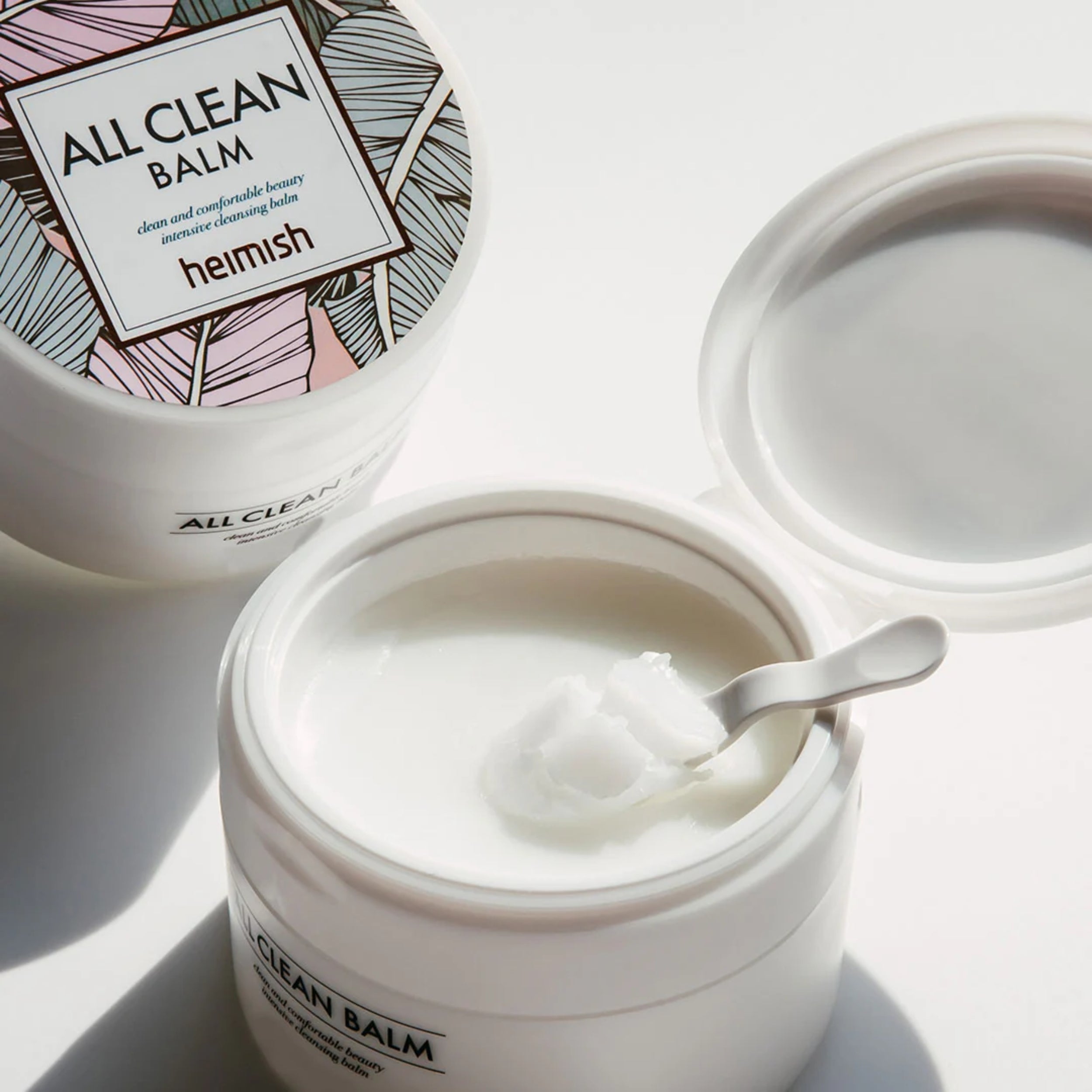Open jar of 'All Clean Balm' with a spoon inside, surrounded by packaging on a light background.