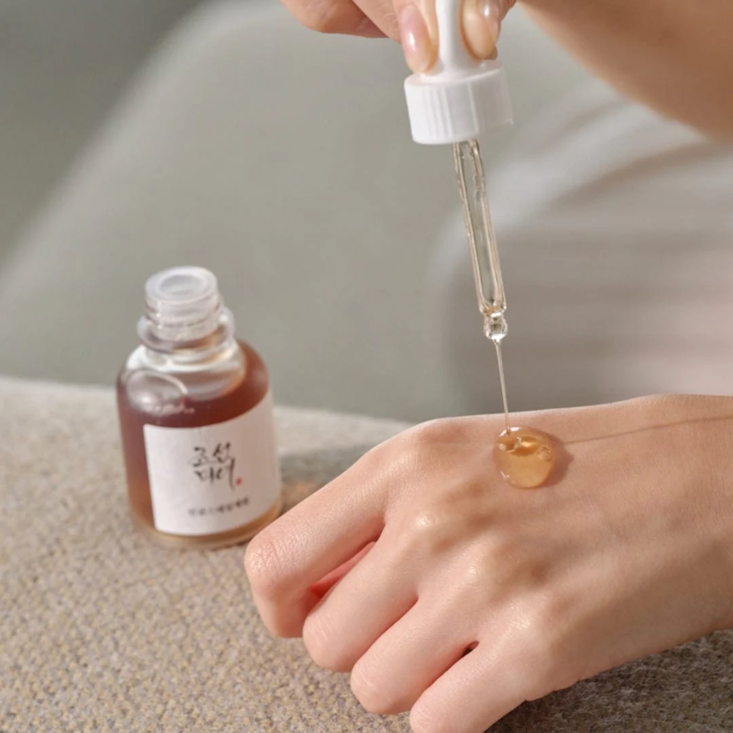 Hand applying a dropper of Revive Serum : Ginseng + Snail Mucin to skin with a bottle labeled 'Klorane' in the background.