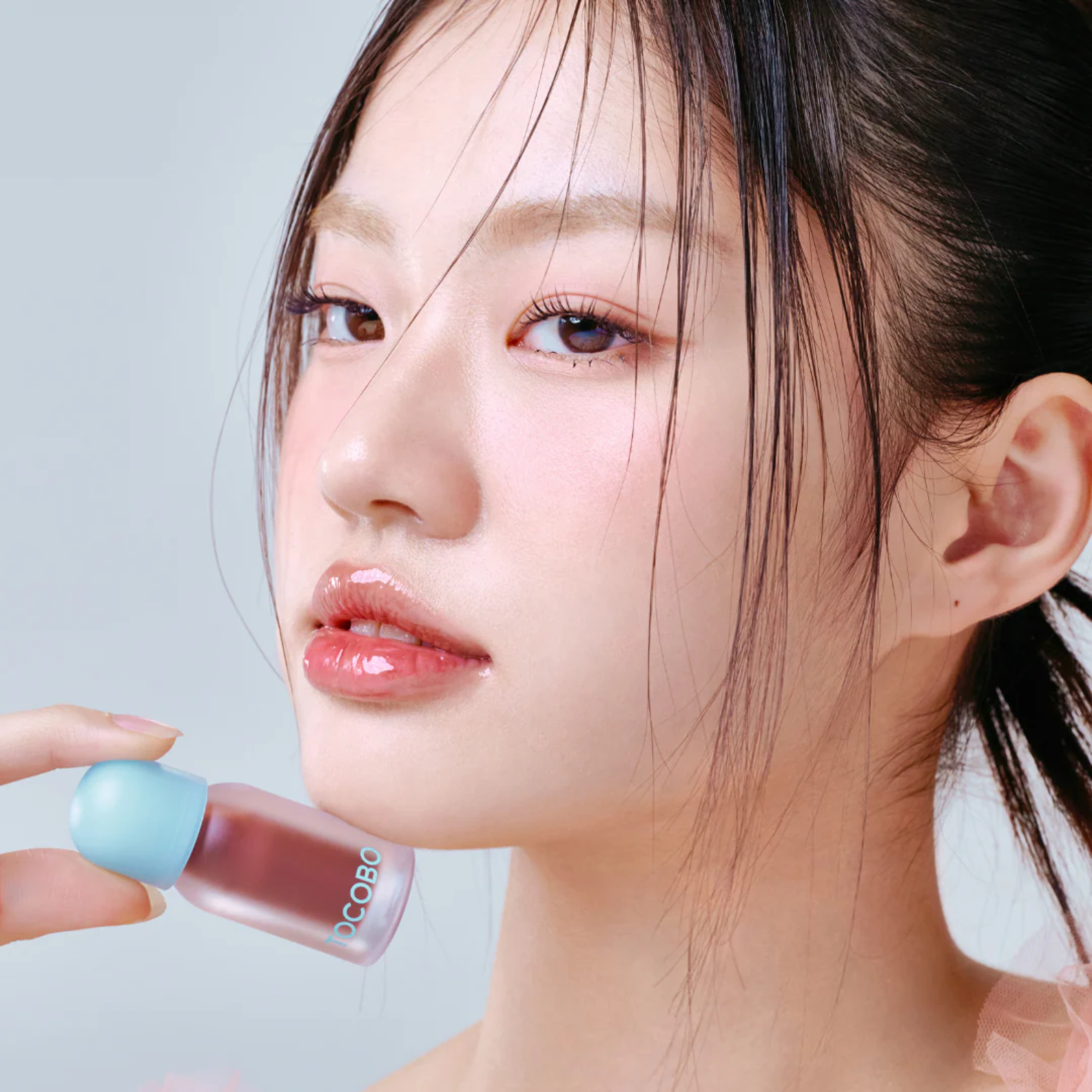 A close-up lifestyle shot of a woman with glossy pink lips holding a small bottle of lip gloss near her mouth. The bottle has a light blue cap and a frosted glass body showing a dusty rose pink liquid inside.